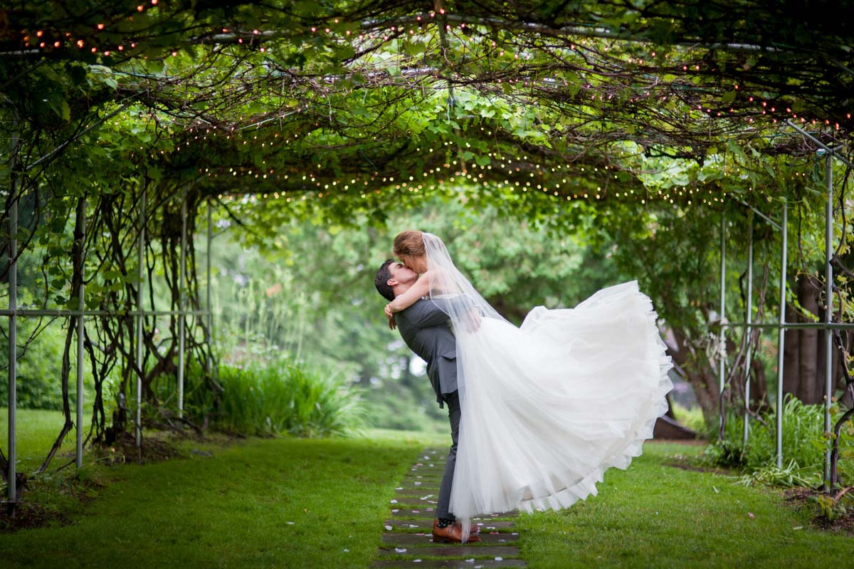 groom lifts bride in a kiss under twinkle lights of grape arbor